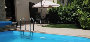 Ein Schwimmbecken mit Metallleiter, eine Terrasse mit Stühlen und Sonnenschirm sowie ein gepflegter Rasen.