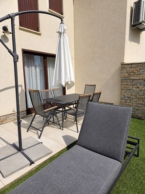 Una terrazza esterna con tavolo e sedie, una sdraio e un pergolato con un ombrellone bianco.