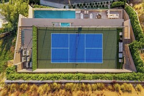 Une vue aérienne d’un court de tennis, avec une piscine à côté, le tout entouré de verdure et d’un bâtiment doté d’une terrasse.