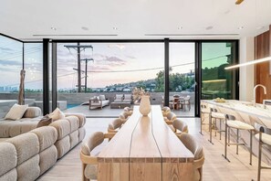 Un espace repas moderne avec une longue table en bois et des chaises, un coin bar avec des tabourets, et de grandes fenêtres offrant une vue sur le paysage urbain.