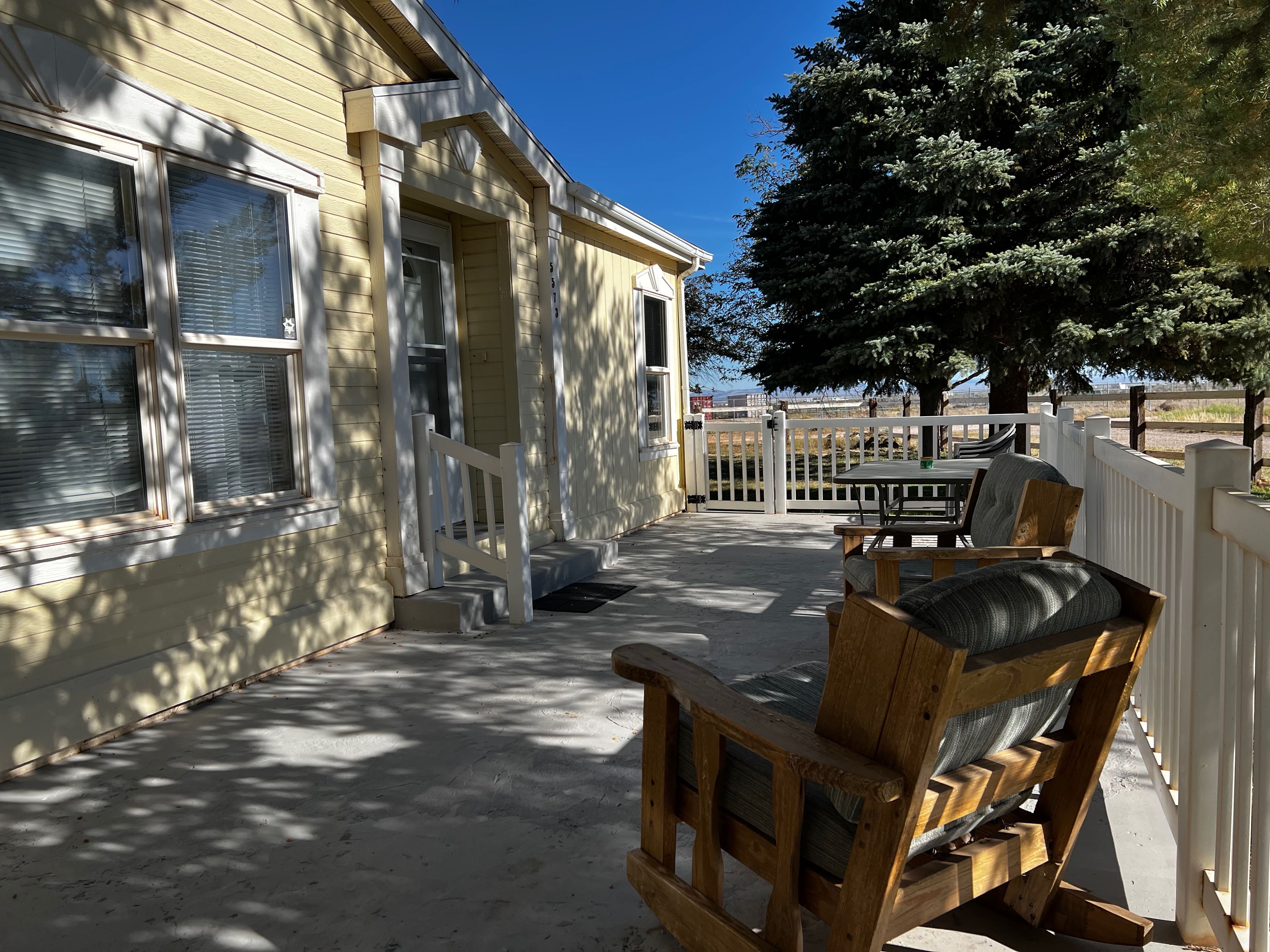 Un patio cubierto con mobiliario exterior, una cerca blanca y un árbol que da sombra.