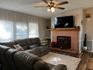 A living room with a leather sofa, a flat-screen TV mounted above a brick fireplace, and a ceiling fan. - Pines Peaks Retreat - 3 bedroom 2 bath, sleeps 8 - Gateway to Southern Utah (Cedar City)
