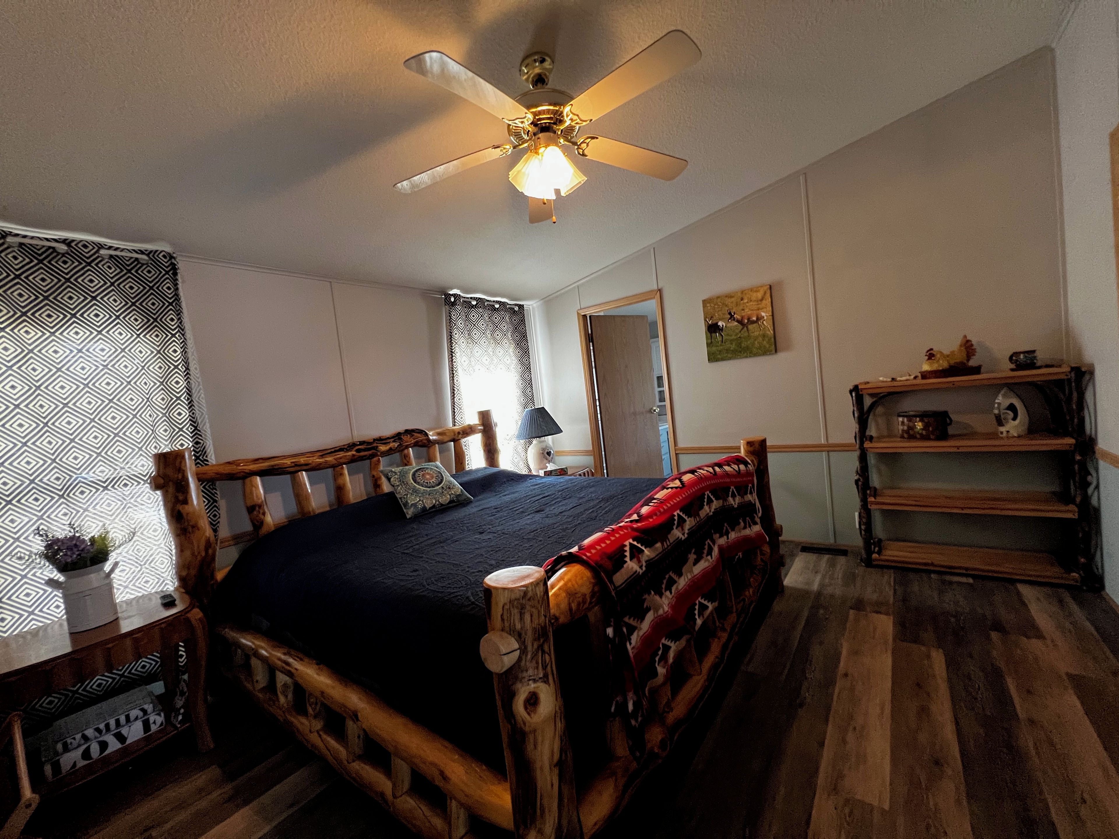 A bedroom with a wooden bed, a ceiling fan, a bedside table, a lamp, a shelf with kitchen items, and a door.