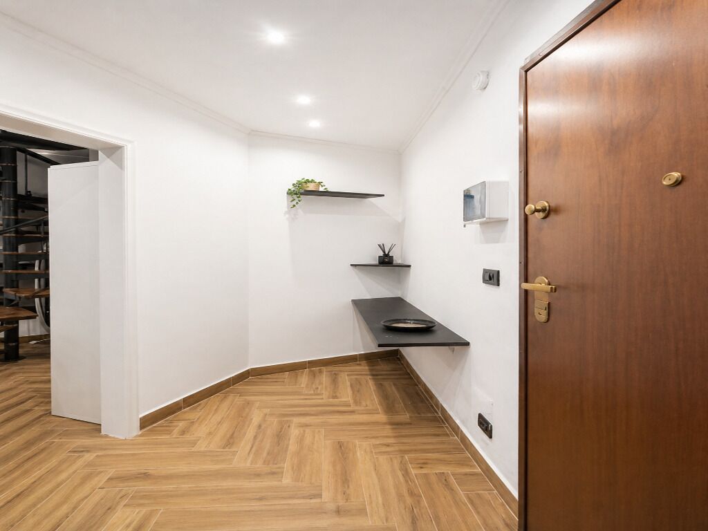 Un pasillo con piso de madera, una estantería negra con una planta y una puerta con manija de bronce.