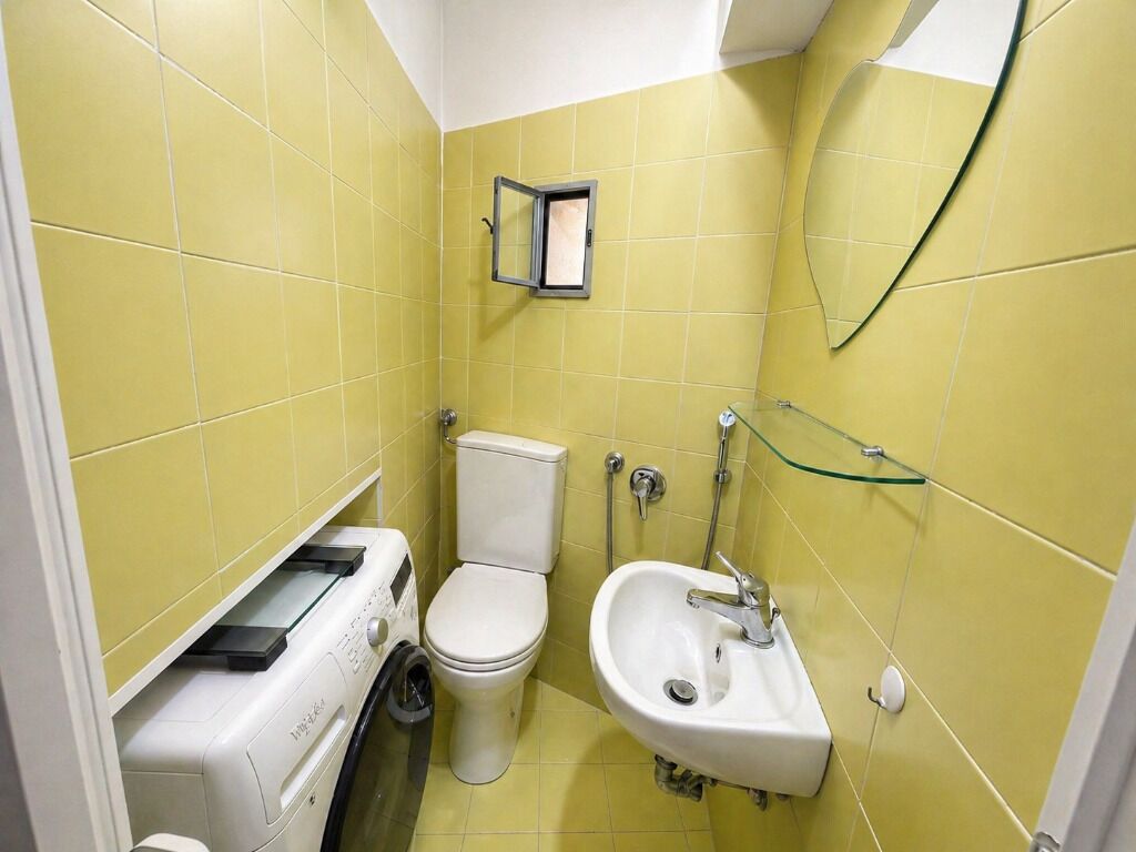 A compact bathroom with yellow tiles, a toilet, a washing machine, and a sink.