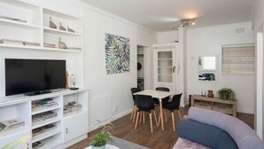Ein modernes Wohnzimmer mit Flachbildfernseher, weißen Regalen, einem Esstisch mit Stühlen und einer Couch.