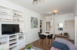 A modern living room with a flat-screen TV, white shelving, a dining table with chairs, and a sofa. - Stylish Gardens Apt Views, Steps from Kloof Street (Cape Town)