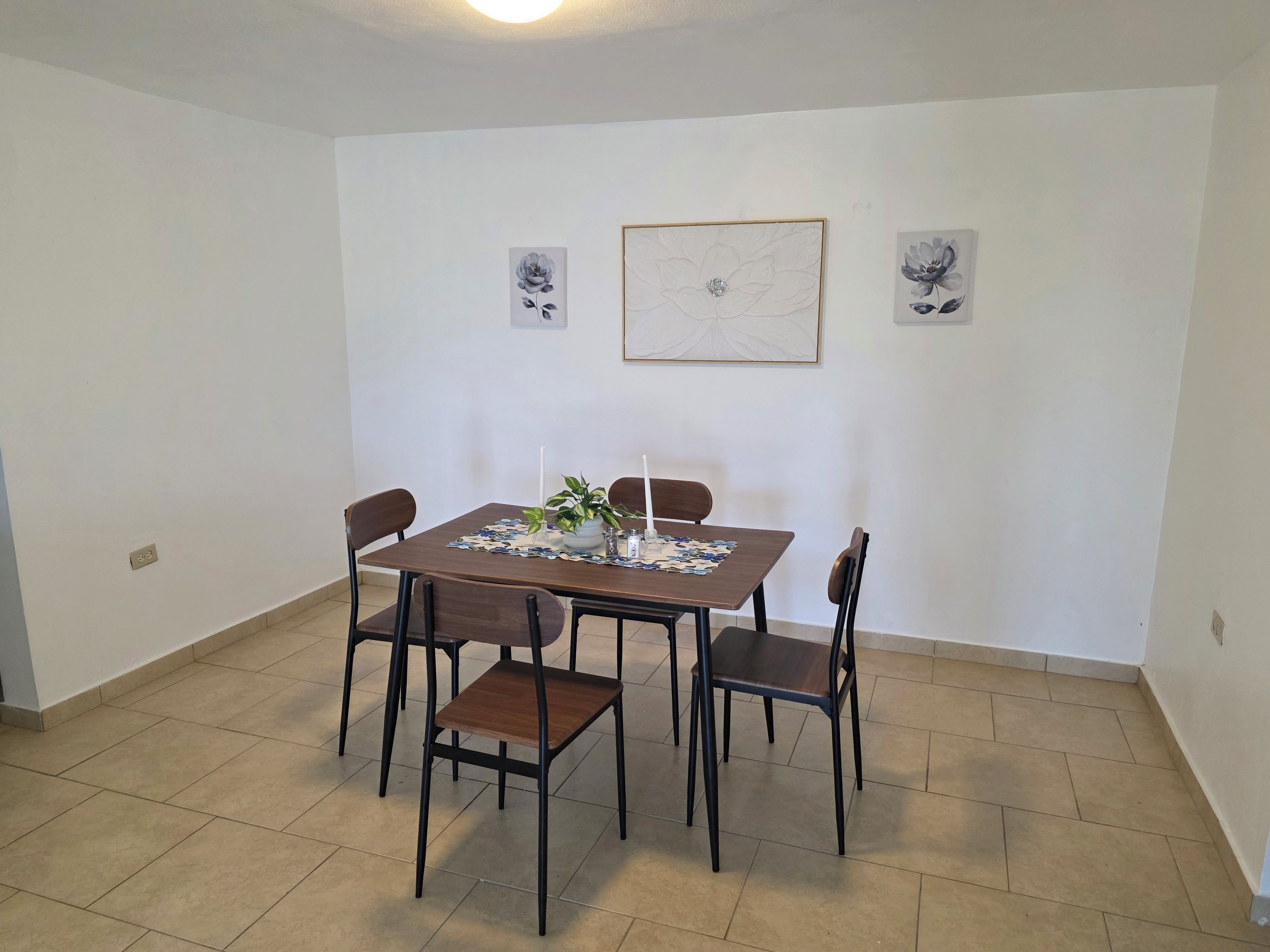 Een eetgedeelte met een vierkante tafel, vier stoelen, een potplant en twee bloemachtige kunstwerken aan de muur.