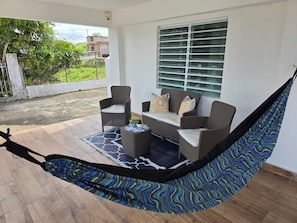 A hammock chair with a patterned fabric, a wicker sofa with cushions, a small table with a plant, and a view of a residential area through a window with blinds.