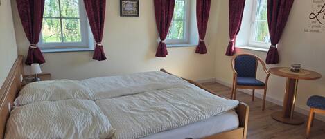 A bedroom with a double bed, a chair, a small table, and a window with curtains.