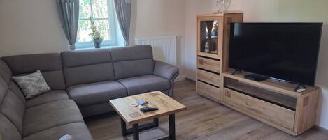 A living room with a sofa, a coffee table, a TV on a wooden stand, and a window with curtains.