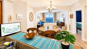 A spacious living room with a dining area, a large window offering a view, and a television displaying 'Dunn Deal Properties & Vacation Rentals'. - Legacy Tower II 802 | Stunning Coastal Views! (Gulfport)