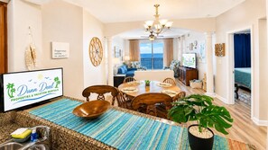 A spacious living room with a dining area, a large window offering a view, and a television displaying 'Dunn Deal Properties & Vacation Rentals'. - Legacy Tower II 802 | Stunning Coastal Views! (Gulfport)