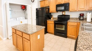 A kitchen with wooden cabinets, a granite countertop, a black refrigerator, a microwave, a stove, and a coffee maker. - Legacy Tower II 802 | Stunning Coastal Views! (Gulfport)