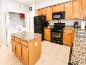 A kitchen with wooden cabinets, a granite countertop, a black refrigerator, a microwave, a stove, and a coffee maker. - Legacy Tower II 802 | Stunning Coastal Views! (Gulfport)