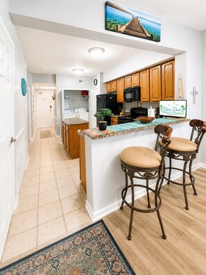 A kitchen with wooden cabinets, a central island with bar stools, a microwave, and a refrigerator. - Legacy Tower II 802 | Stunning Coastal Views! (Gulfport)