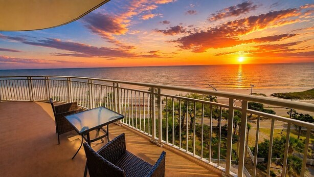 Varanda com mesa e cadeiras, com vista para o pôr do sol sobre o oceano.