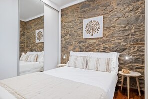 A bedroom with a stone wall, a bed with white bedding, a bedside table with a lamp, and a mirror reflecting the room. - Stone House Rochinha by Homie (Funchal)