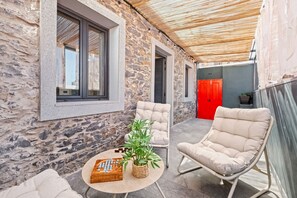 A stone-walled outdoor seating area with two cushioned chairs, a small round table with a plant and a wooden box, and a red door in the background. - Stone House Rochinha by Homie (Funchal)