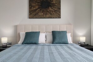 Ein ordentlich bezogenes Bett mit einem blauen, gemusterten Bettdeckenüberwurf, zwei Kissen und einem an der Wand hängenden, abstrakten Kunstwerk.