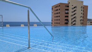 A swimming pool with clear blue water, a metal pool ladder, and a view of the ocean and a tall building in the background. - SUNSET SAILORS cozy apartment (Benidorm)