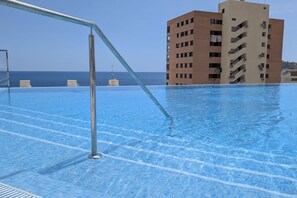 Ein Schwimmbecken mit klarem, blauem Wasser, einer Metallleiter und Blick auf den Ozean sowie ein hohes Gebäude im Hintergrund.