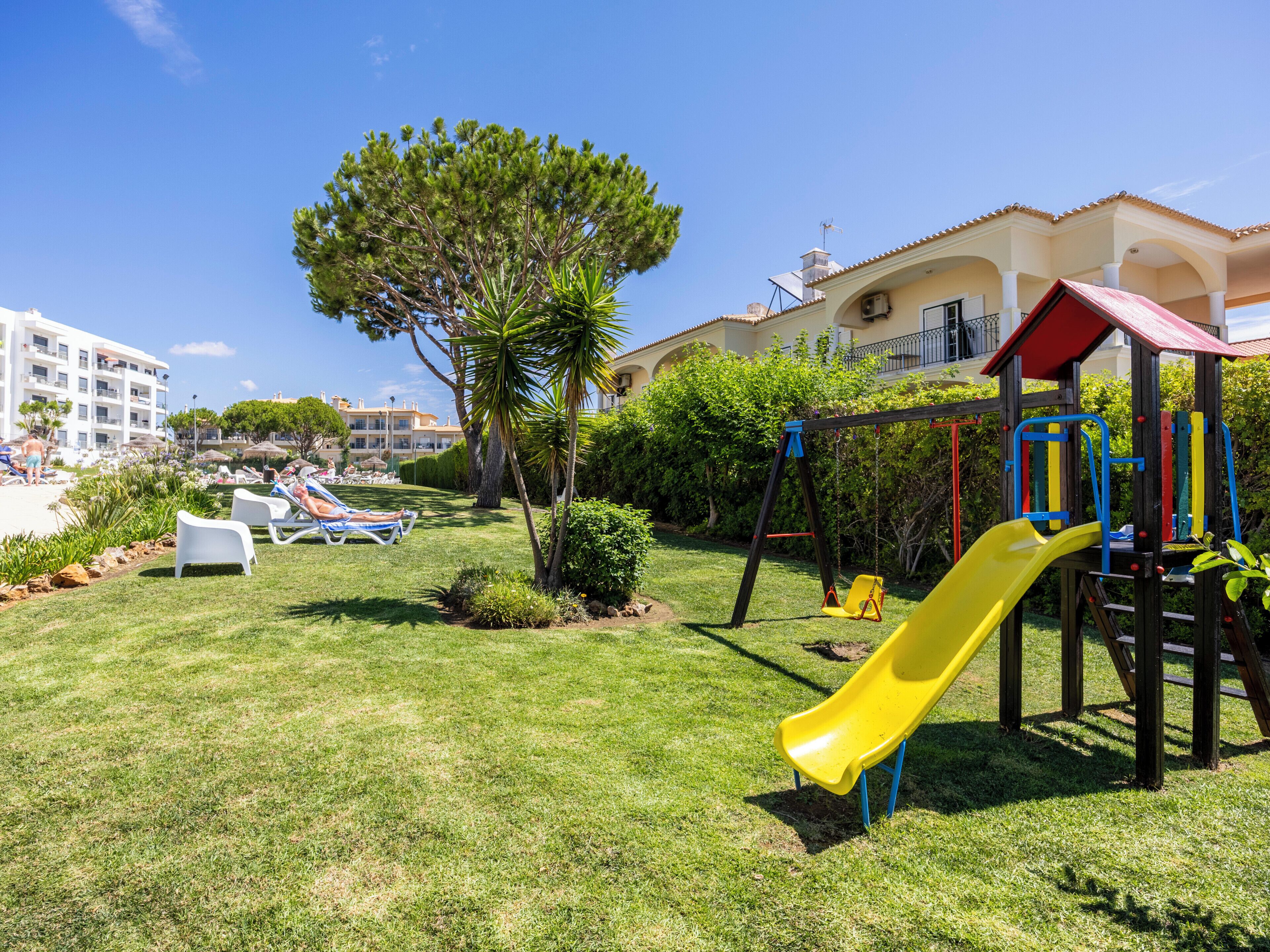A playground with a yellow slide, surrounded by grass and trees, with a building in the background.
