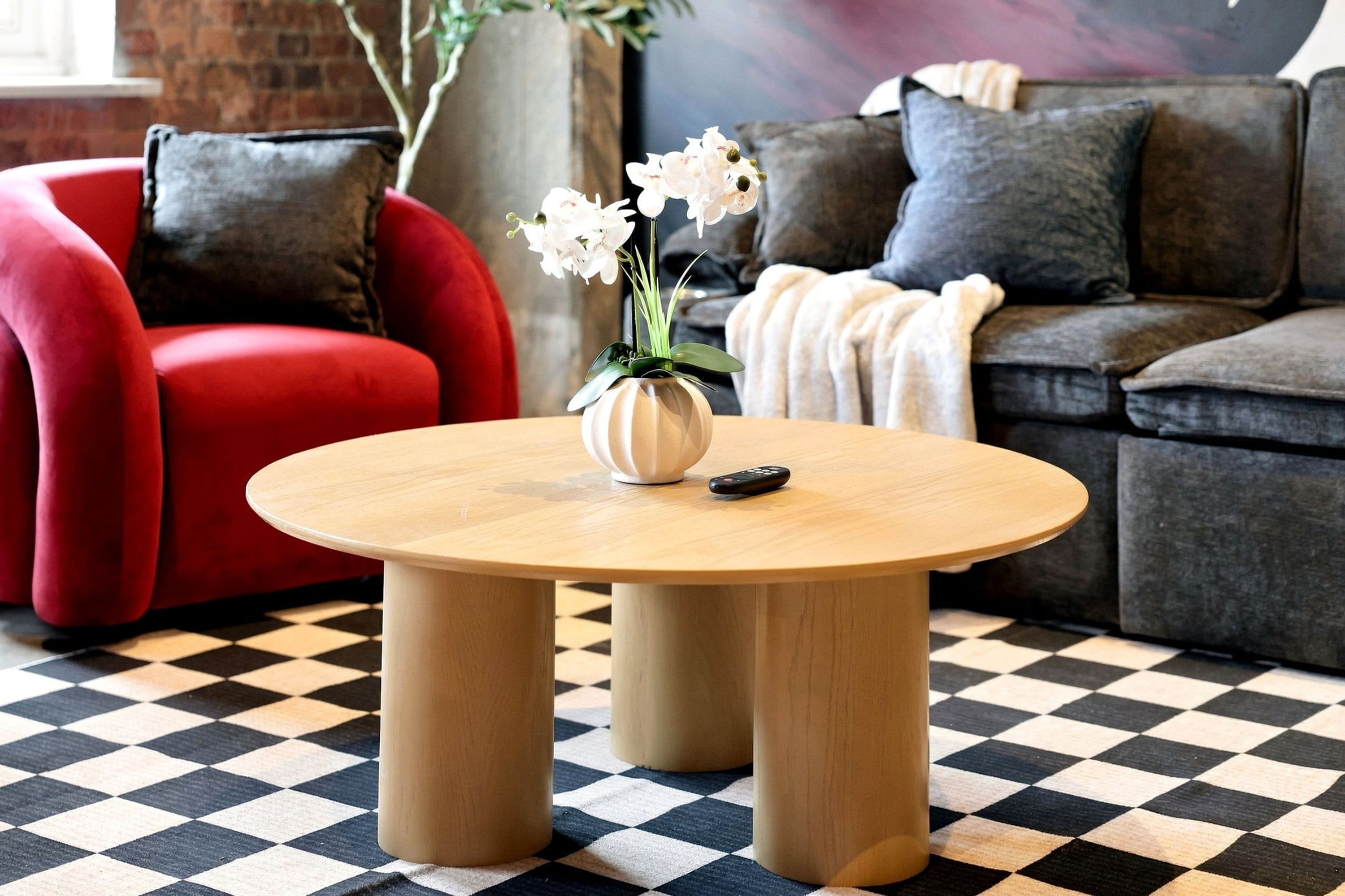 A round wooden table with a vase of white flowers and a remote control on it, placed in a room with a checkered floor, red and grey sofas, and a brick wall.