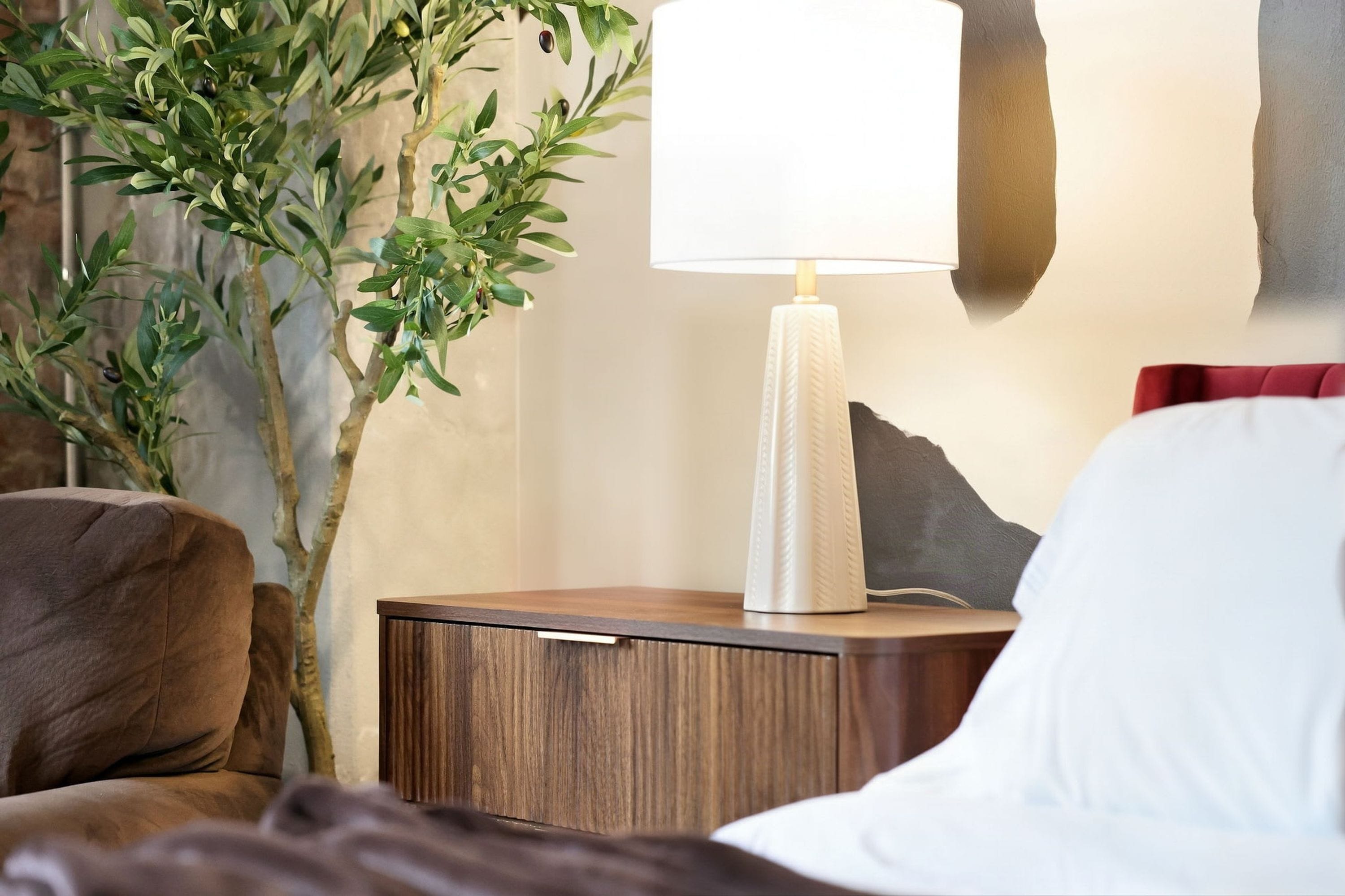 A bedroom with a wooden nightstand, a white lamp, a red pillow, and a green plant.