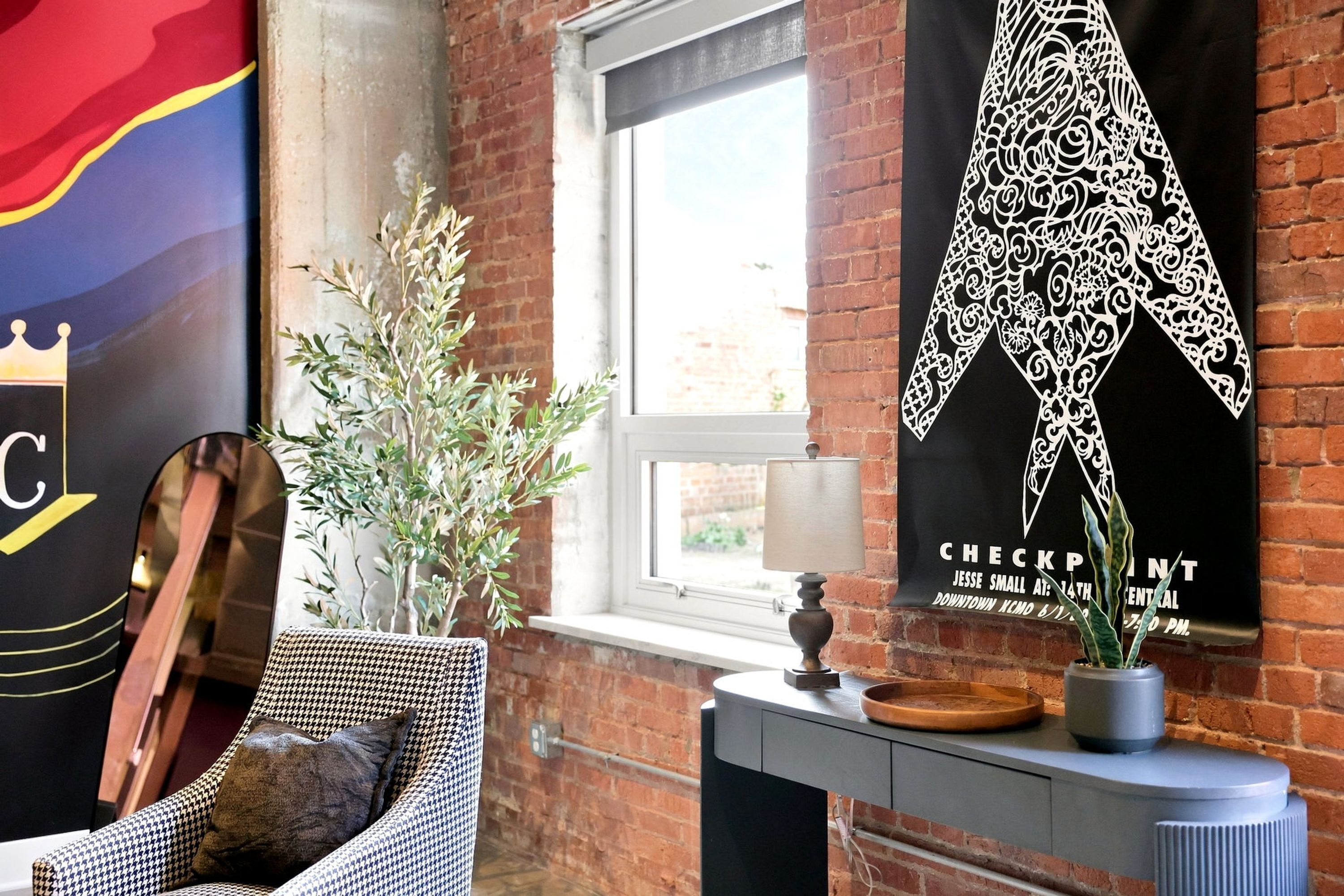 A room with a houndstooth patterned armchair, a potted plant, a wall-mounted lamp, and a poster advertising a check-in event.