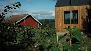 Une grange rouge et une structure moderne en bois dotée d’une grande fenêtre, le tout entouré d’une végétation luxuriante.