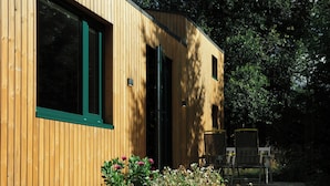 Un bâtiment en bois doté de grandes fenêtres et d’une porte, entouré de verdure et de plantes en pot.