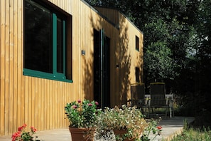 A wooden building with large windows and a door, surrounded by greenery and potted plants.