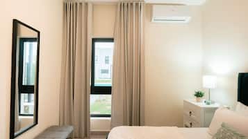 A hotel room with a bed, a bench, a bedside table with a lamp, and a window with curtains.