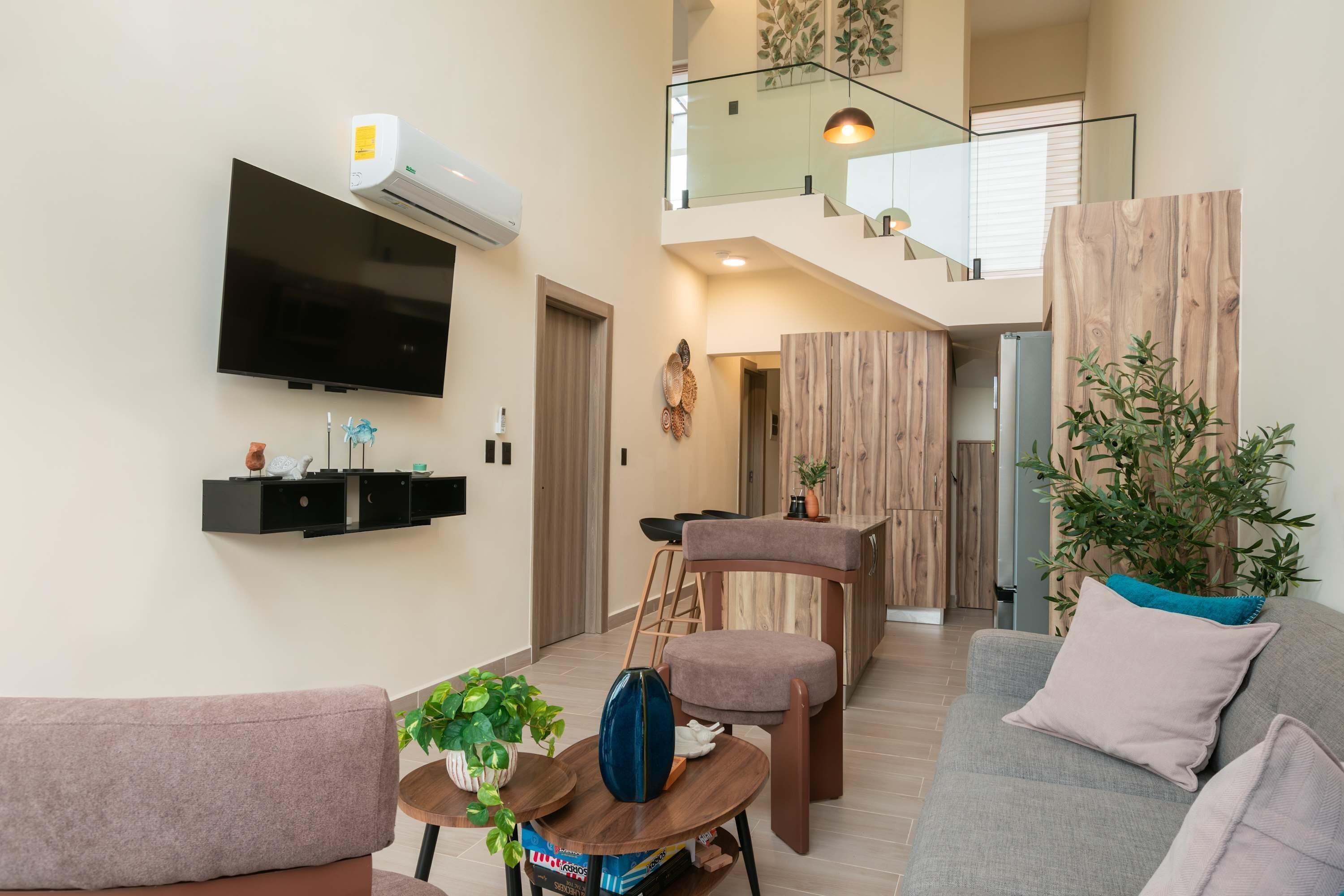 A modern living room with a flat-screen TV, built-in shelves, a dining area with a wooden table, and a staircase leading to an upper level.