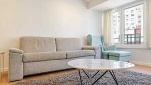 Un salon moderne avec un canapé gris, une table basse ronde et une vue sur un immeuble par la fenêtre.