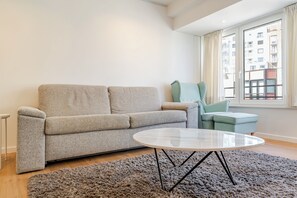 Ein modernes Wohnzimmer mit einem grauen Sofa, einem runden Couchtisch und Blick auf ein Gebäude durch ein Fenster.