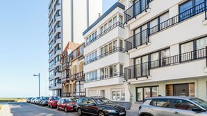 Une rangée d’immeubles résidentiels modernes à plusieurs étages, dotés de balcons, et un espace de stationnement où sont garées différentes voitures.