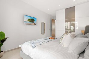 Un dormitorio moderno con una cama, una mesita de noche con frutero, un espejo grande y un cuadro de una playa.