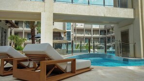 Zona de piscina con sillones de descanso y barandilla de cristal con vistas a un edificio con balcones.
