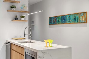 A modern kitchen with a white marble countertop, a stainless steel faucet, and a built-in oven. There are wooden shelves with plants and a colorful abstract painting on the wall. - 5 Minutes to Eagle Beach|Private BBQ| Pool & Jacuzzi (Noord)