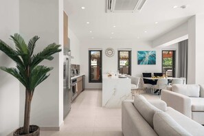 Eine moderne Küche mit einer zentralen Kochinsel, ein Essbereich mit weißem Tisch und Stühlen sowie ein Wohnbereich mit einer Couch und einer großen Pflanze.