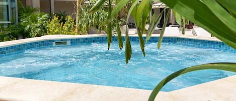 Une piscine dont le bord est recouvert de carreaux, entourée de rochers et de plantes, avec un immeuble à plusieurs étages en arrière-plan.