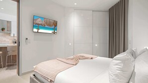 A modern bedroom with a bed, a TV mounted on the wall, and a bathroom with a shower. - Breezy Balcony | Top-Floor 1BR | Near Eagle Beach (Noord)