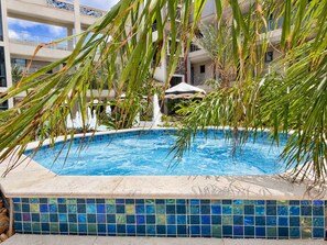 A swimming pool with a tiled edge, surrounded by palm trees and a building with balconies in the background. - Breezy Balcony | Top-Floor 1BR | Near Eagle Beach (Noord)