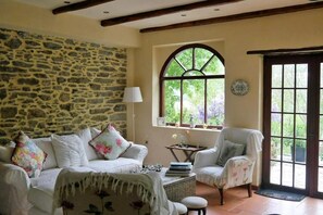 Una habitación acogedora con pared de piedra, un sofá blanco con cojines florales, una mesita con libros y una silla.