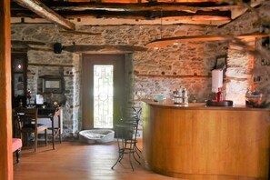 A rustic interior with a stone wall, wooden ceiling beams, a bar area with stools, and a dining table with chairs.