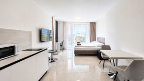 Moderni hotellihuone, jossa on pieni keittiö, sänky, ruokailutila ja suuri ikkuna, josta avautuu näkymä kaupunkiin.