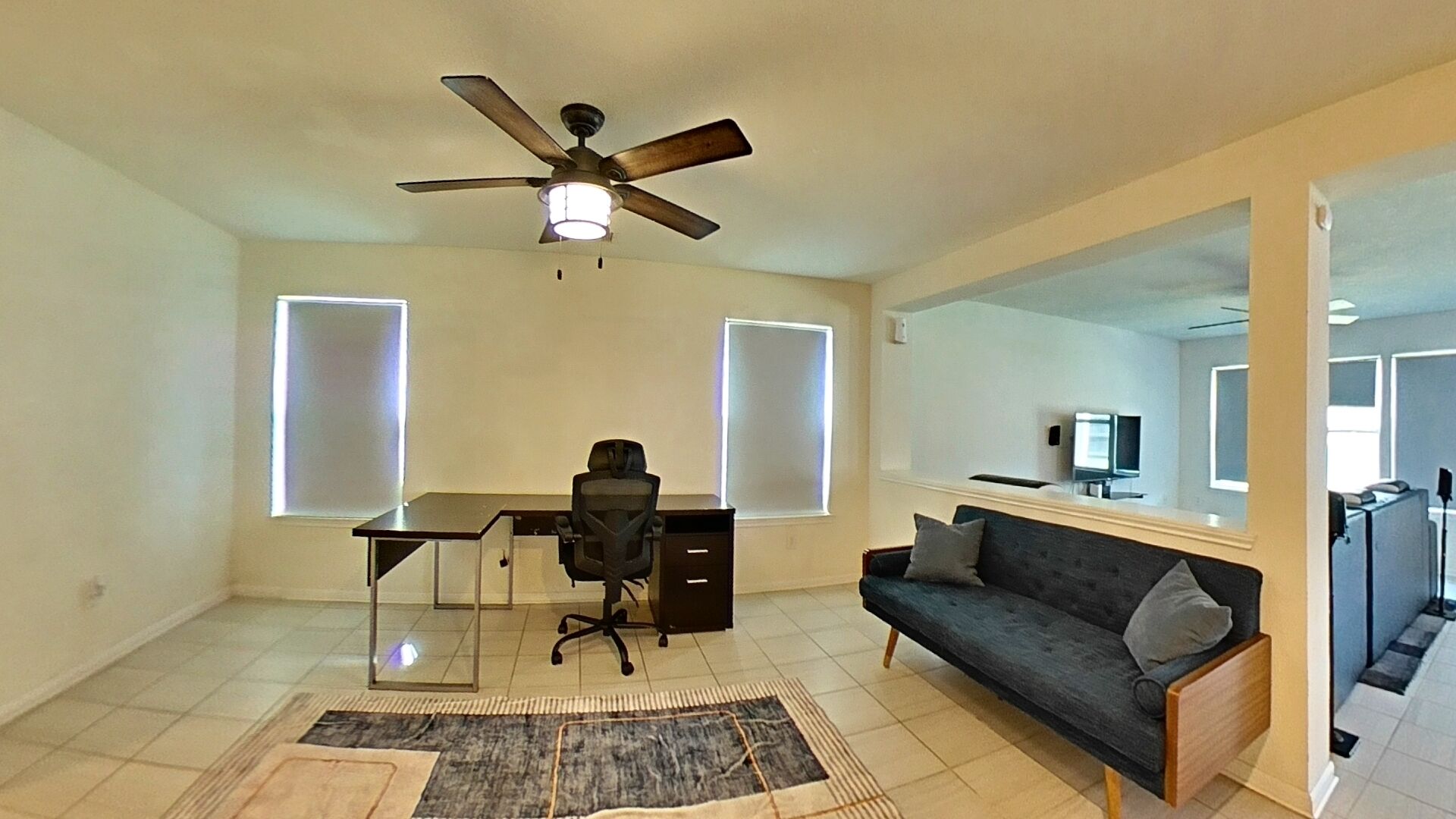 A spacious room with a desk, office chair, and a dark-colored sofa. A ceiling fan is installed above the desk, and a large mirror is mounted on the wall.