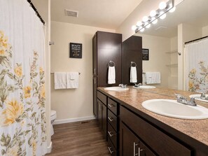 A bathroom with two sinks, a mirror with lights, and a shower curtain with floral patterns. - Unwind at Midtown Haven (Coeur d'Alene)